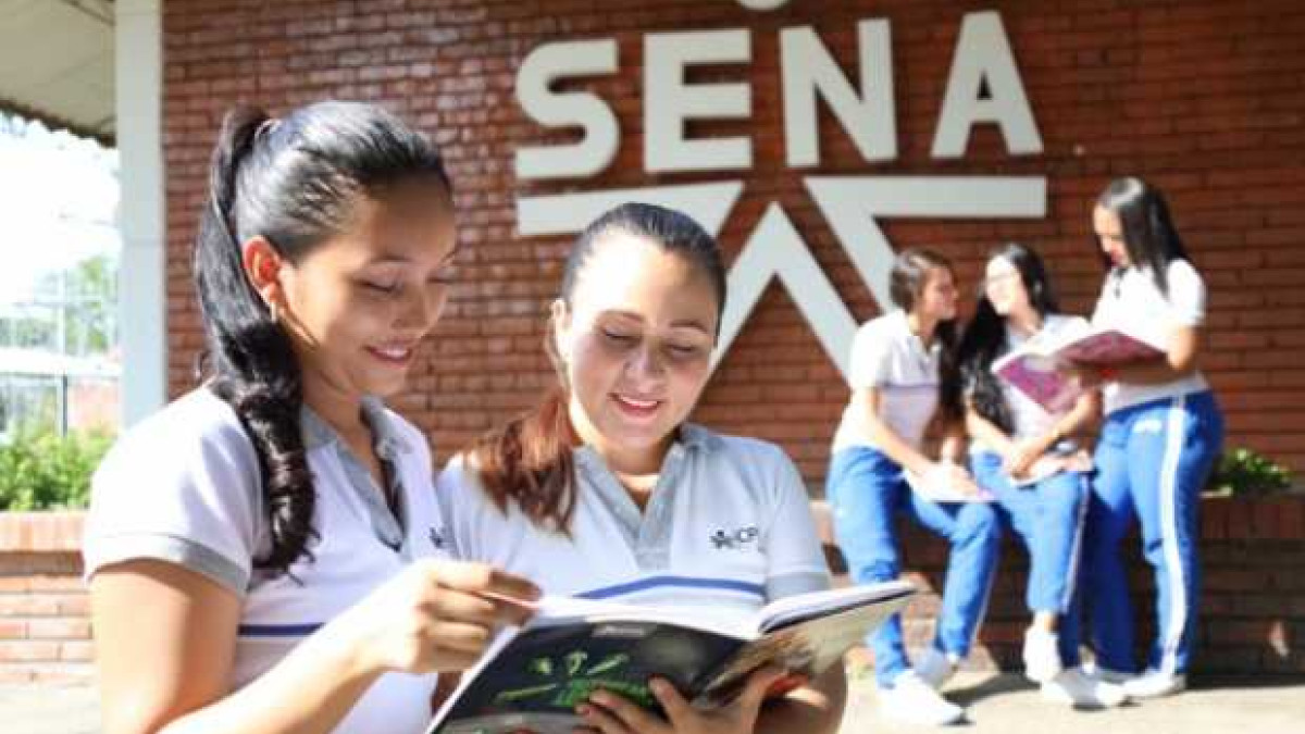 Dos estudiantes del SENA mirando un cuaderno con una pared con el logo del SENA en el fondo.