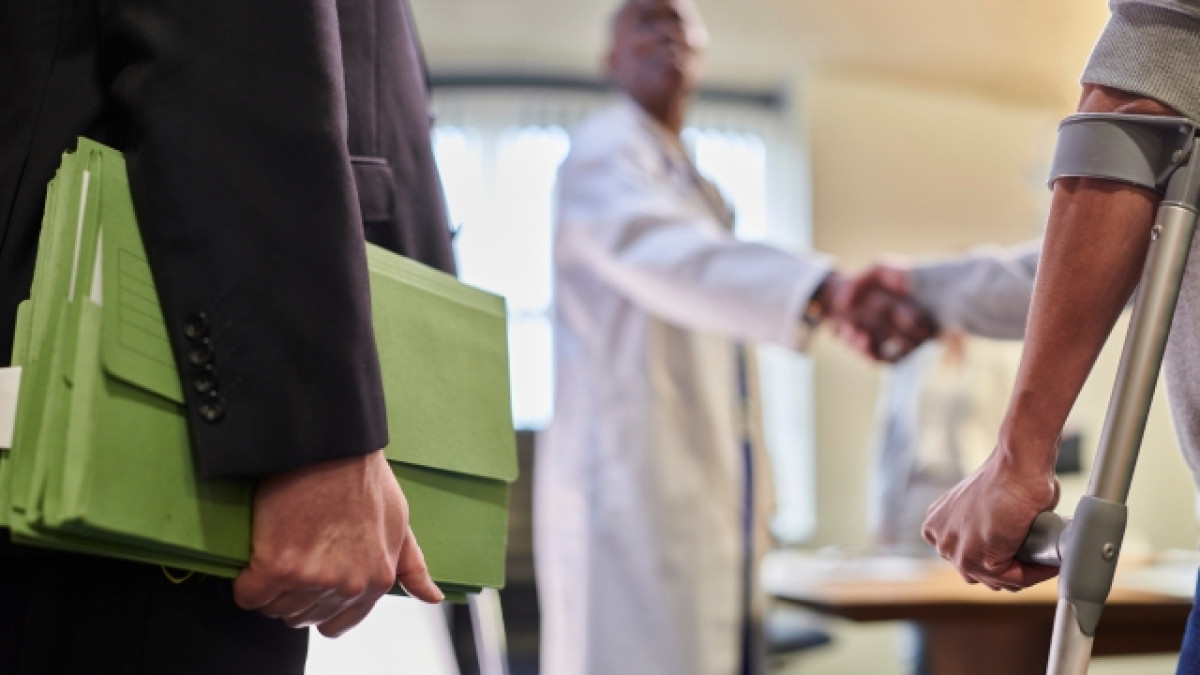 Medico saluda a un hombre en muletas con una persona al lado sosteniendo una carpeta verde.