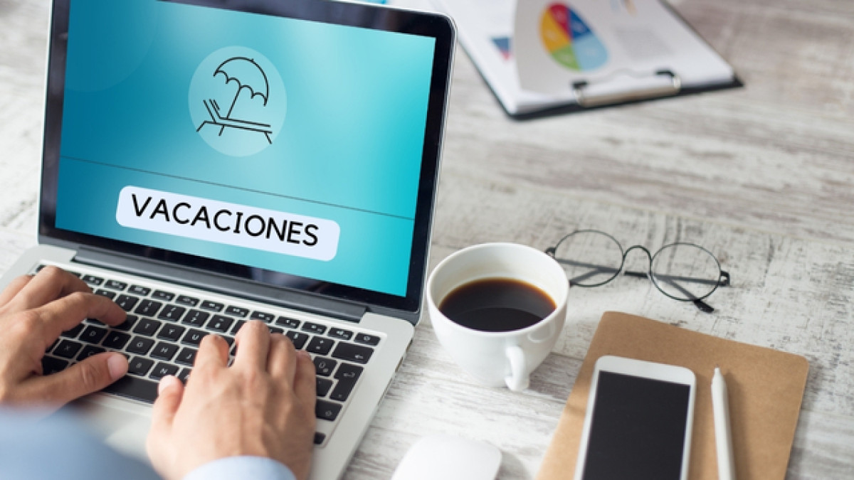 Persona teclea en un computador que dice "VACACIONES". Al lado hay un café, un celular y una carpeta.